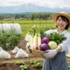収穫したばかりの新鮮な野菜をバスケットに入れて笑顔を見せる日本人女性。背景には、雪解け直後の畑と遠くに見える北海道の山々が広がる。